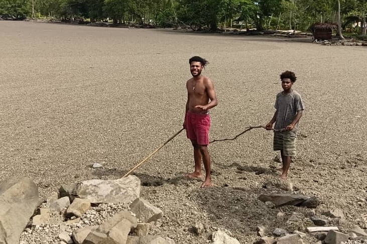Two people with sticks walk along a coastline with stone floating on the ocean behind them