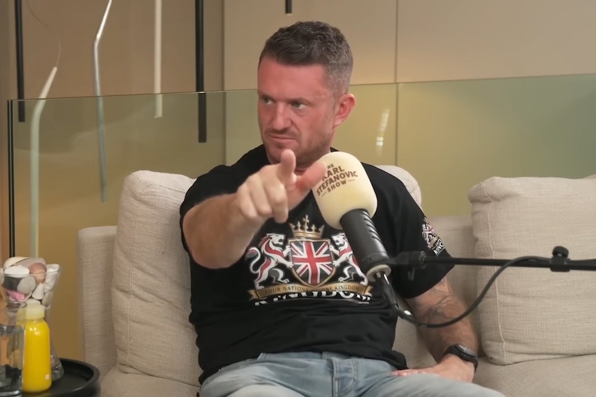 Tommy Robinson, wearing a black shirt, sits behind a microphone, points towards the camera.