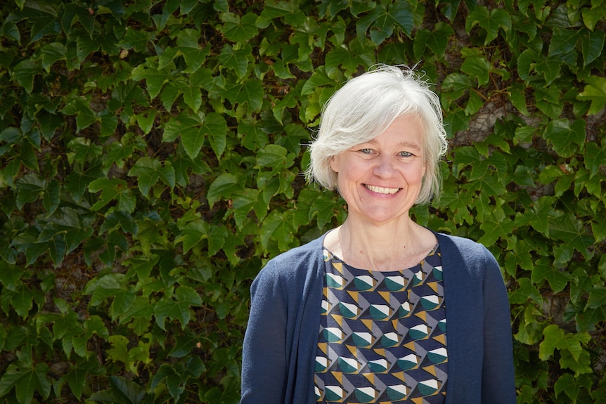 A woman with light white hair and a multicoloured dress and cardigan smiles at the camera.