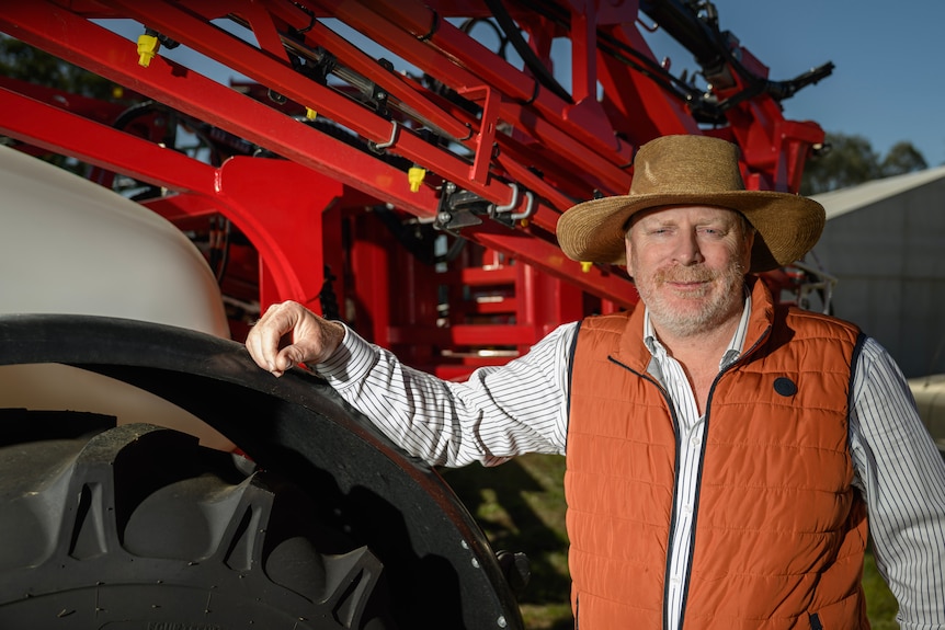 A farmer in an orange vest resting his hand on a large tractor.