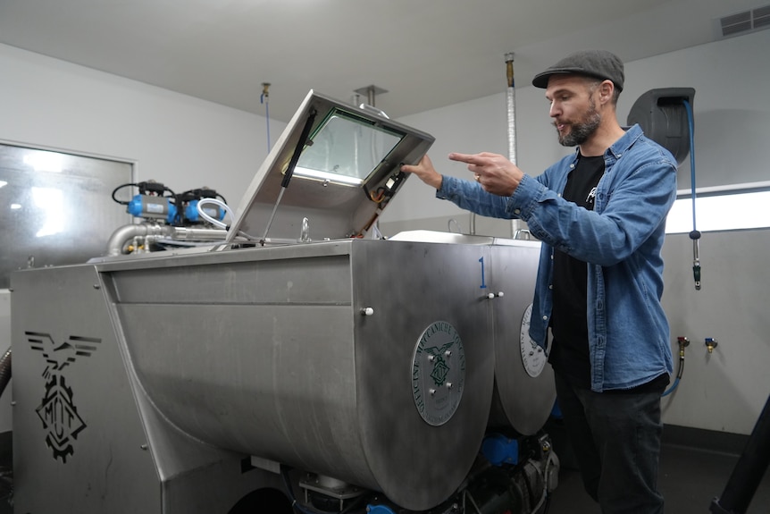 A man in a blue button up shirt and a flat cap points at a olive processing machine.