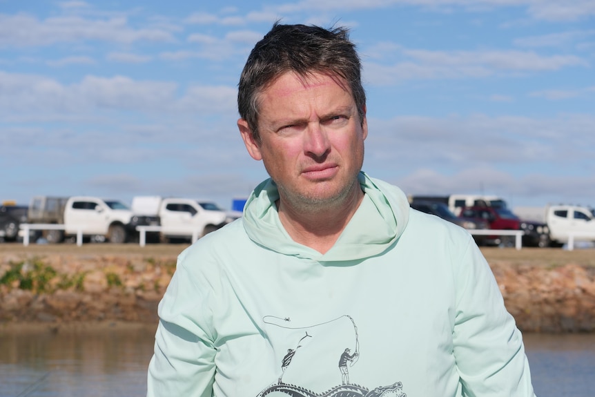 A dark-haired man in a light hoodie stands in front of a waterway on a sunny day.