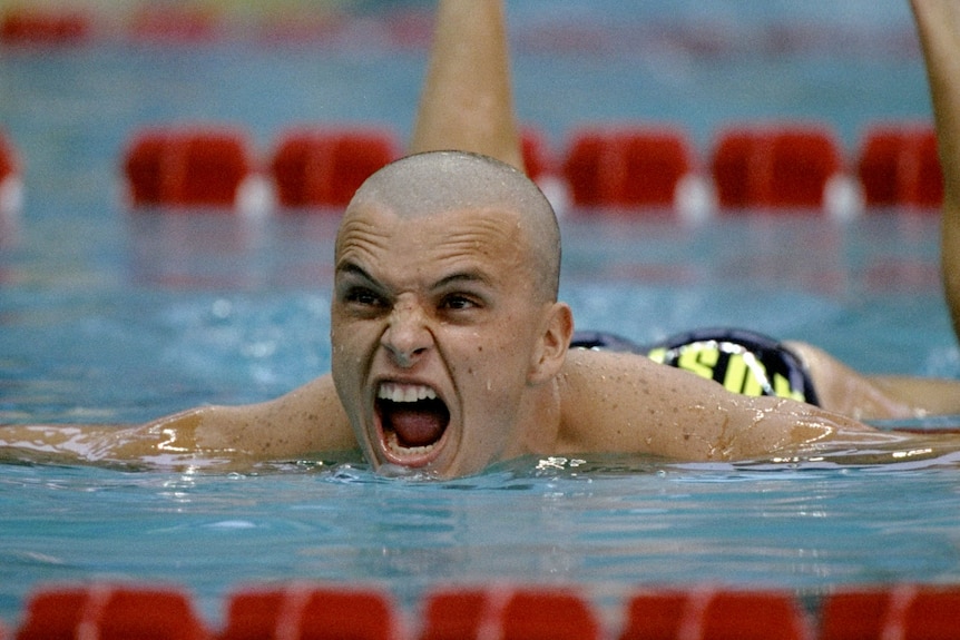 A bald man in a swimming pool yells while swimming over the red lane ropes.