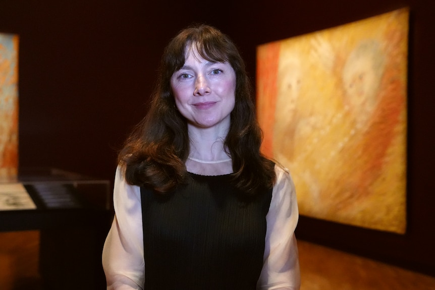 A woman with long drak hair stands in an art gallery smiling lightly.