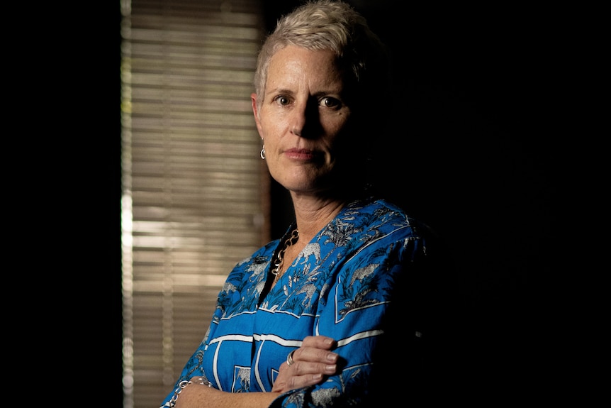A woman with short blond hair stands with her arms crossed looking into the camera with a neutral expression on her face.