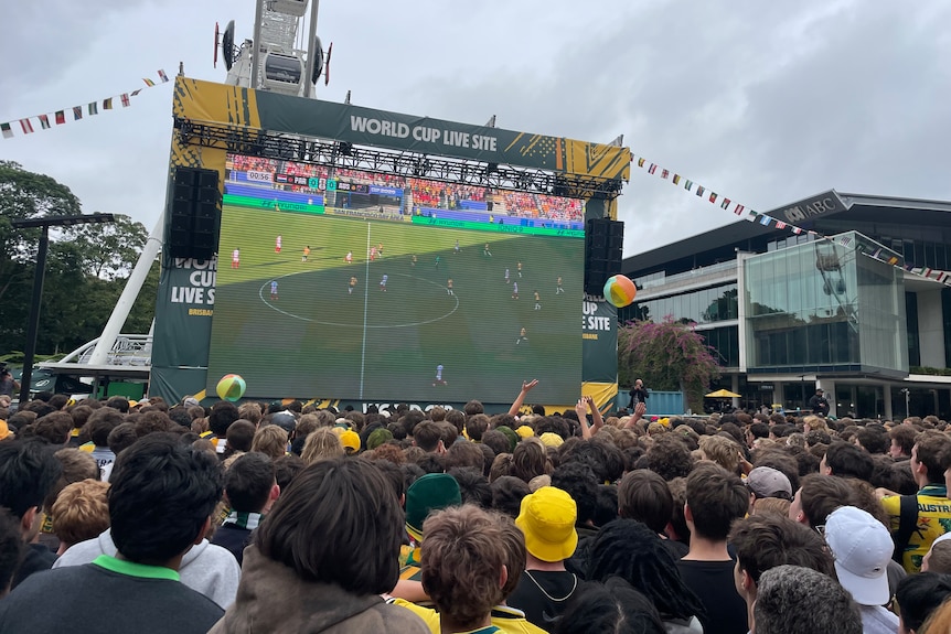 A large screen playing the live soccer game is set up in a public area with hundreds of people standing and watching.
