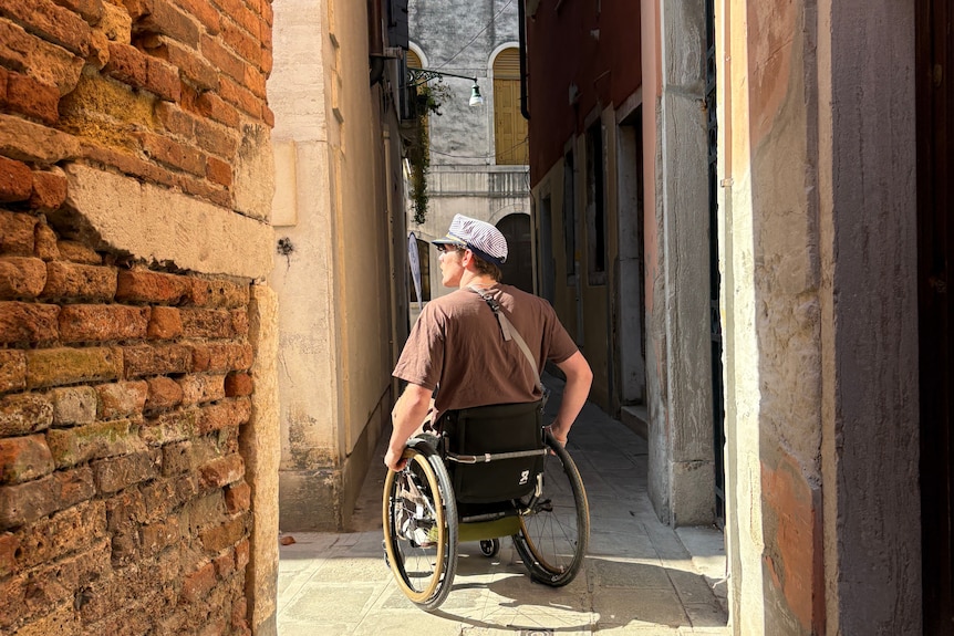 The back of a man in wheelchair in a narrow laneway.