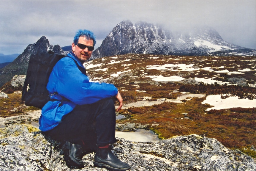 Vintage photo of man with bright blue hair squatting down in front of an epic snowcapped mountain