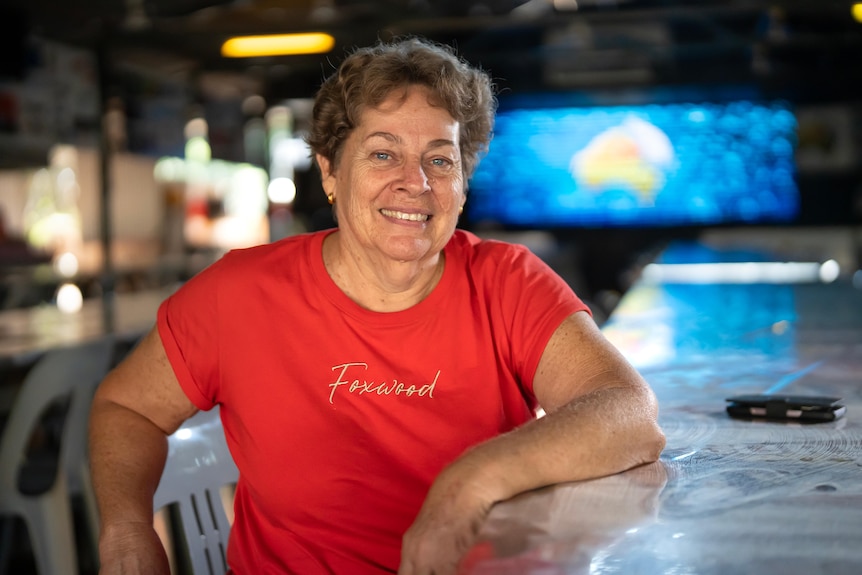 White woman, brown short-cut wavy hair, red-tshirt, sitting at a bar, blue light feature behidn her, yellow map of Australia