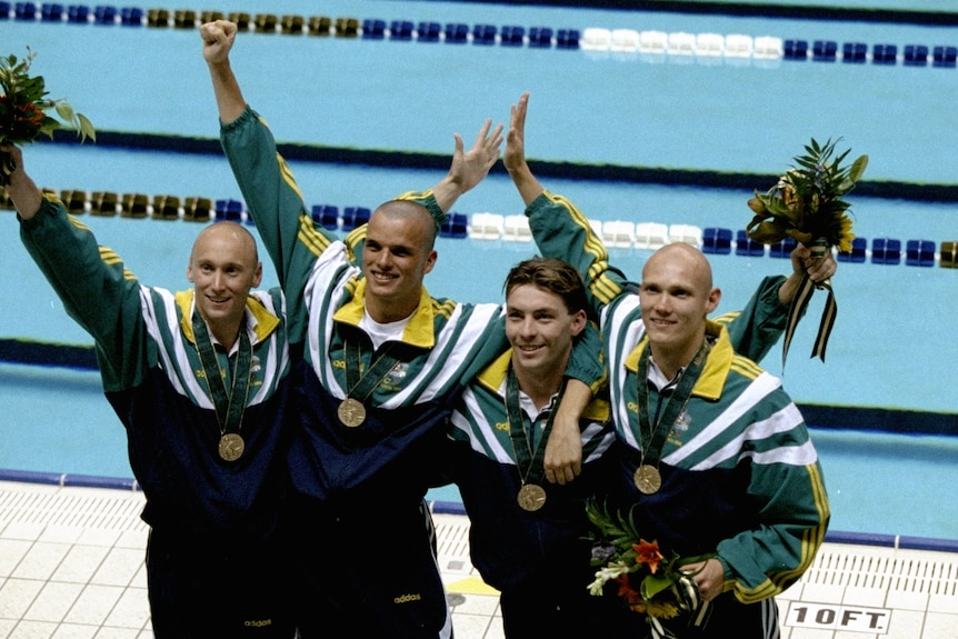 Four men wearing green and gold tracksuits stand in front of a pool with bronze medals around their neck.
