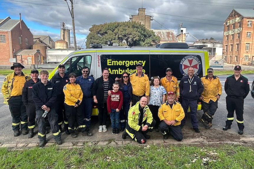 A group of paramedics and firefighters stand in front of an ambulance with a young boy and his family.