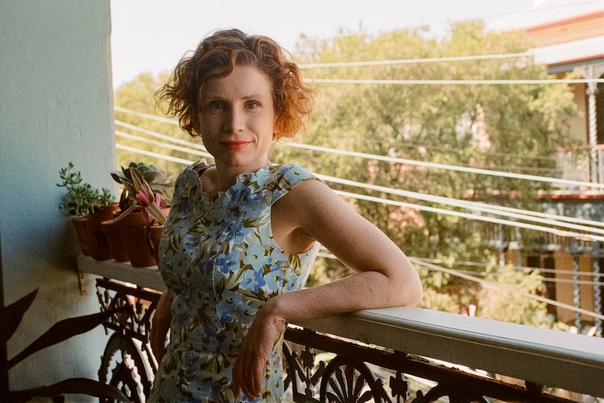 Fiona Wright, late 30s, short read curly hair, smiles slightly, leaning back against the balcony railing of a terrace house.
