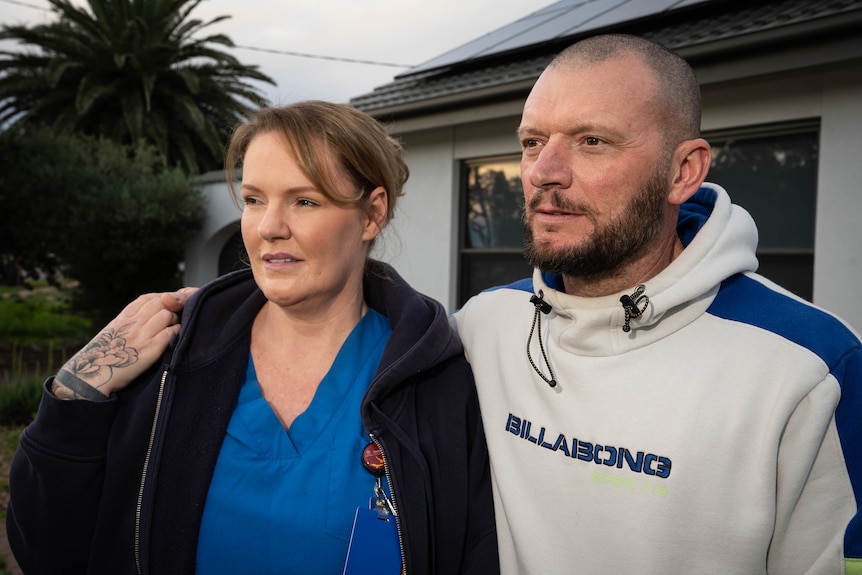 A British midwife with brown hair stands next to her husband in front of their home in Whyalla.