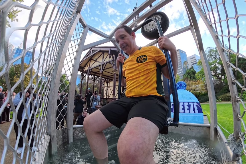 A dark haired man in a green and gold top and green shorts sits on a seat with his legs dangling in water inside a cage.