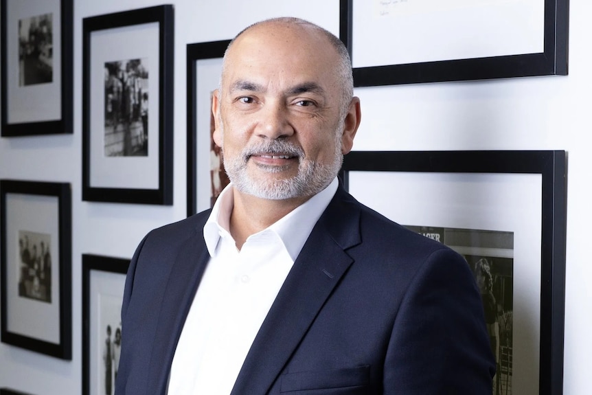 A man wearing a business suit poses professionally in front of a wall of indistinct, black-and-white photographs.