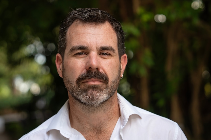 A man with dark brown hair, eyes and beard, moustache, wearing a white collared shirt, serious expression on his face.