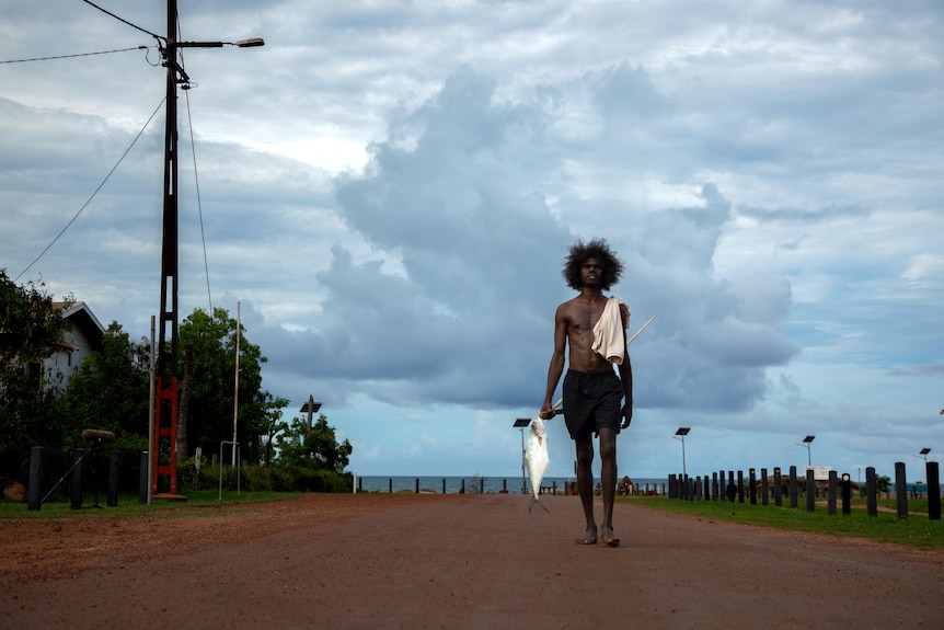 A young Indigenous man with frizzy hair walks along a road shirtless, holding a speared fish by his side.