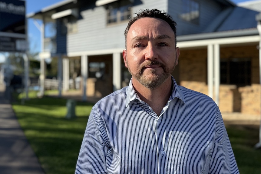 A man with short dark hair, wearing a business shirt, standing outside a house.