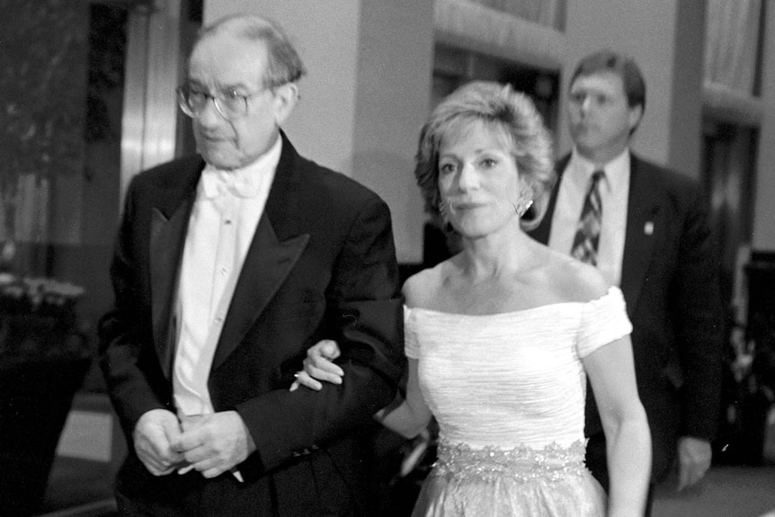 A black and white photo of Alan Greenspan in a tuxedo with his arm linked around Andrea Mitchell wearing a dress.