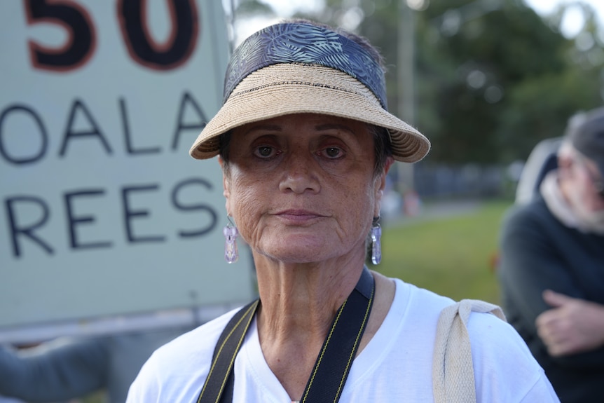 A tan-skinned woman in a wide-brimmed hat poses for camera. She is standing in front of a placard being held up by someone.
