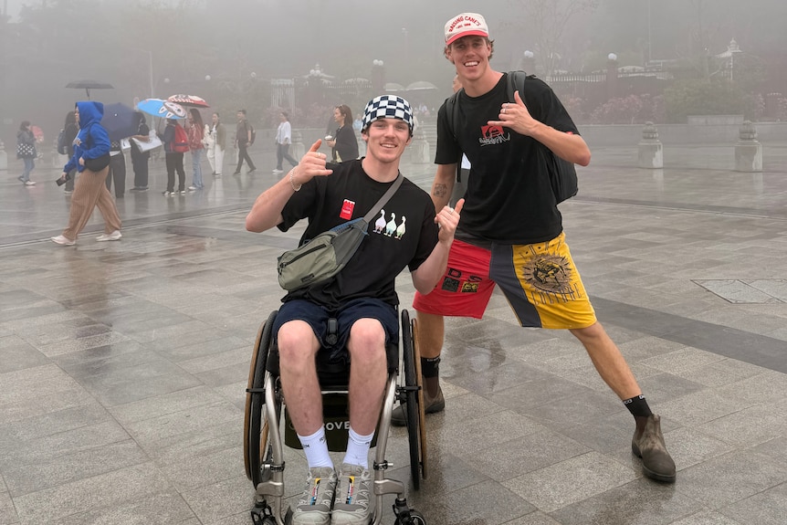 Two men, one in a wheelchair, smiling with thumbs up on a misty day.