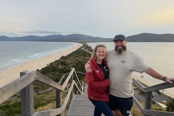 A man and woman embrace while smiling at the camera standing on wooden stairs going down to a beach.