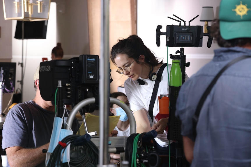 Zoe Pepper, late 30s, glasses, hair tied back, headphones around neck, concentrates, while working on set, cameras around her.