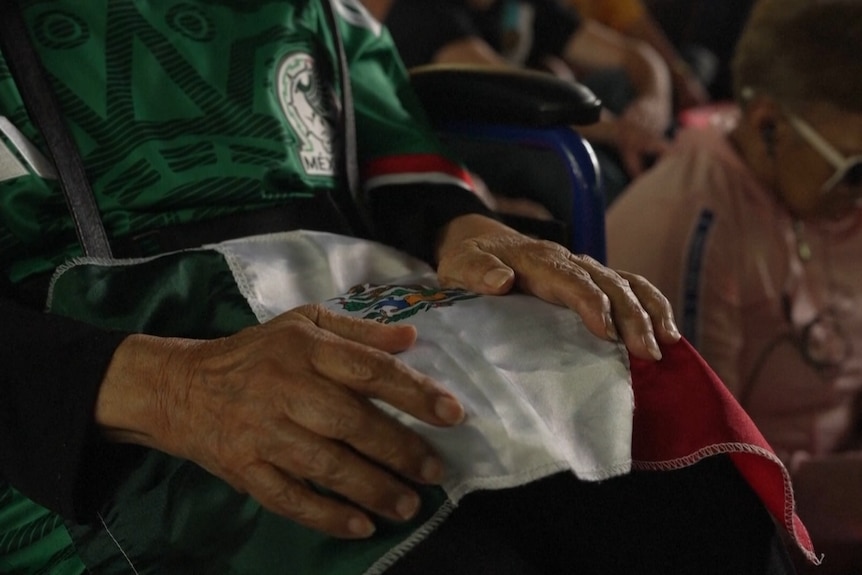a Mexican flag draped across a lap, while two eldery hands rest on top