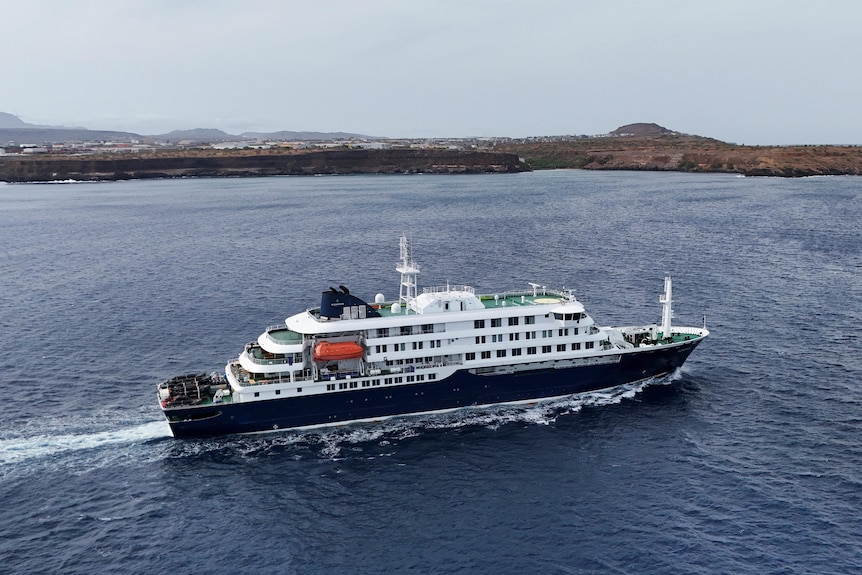 A drone shot shows a cruise ship travelling to the right of screen, in front of a nearby coastline.