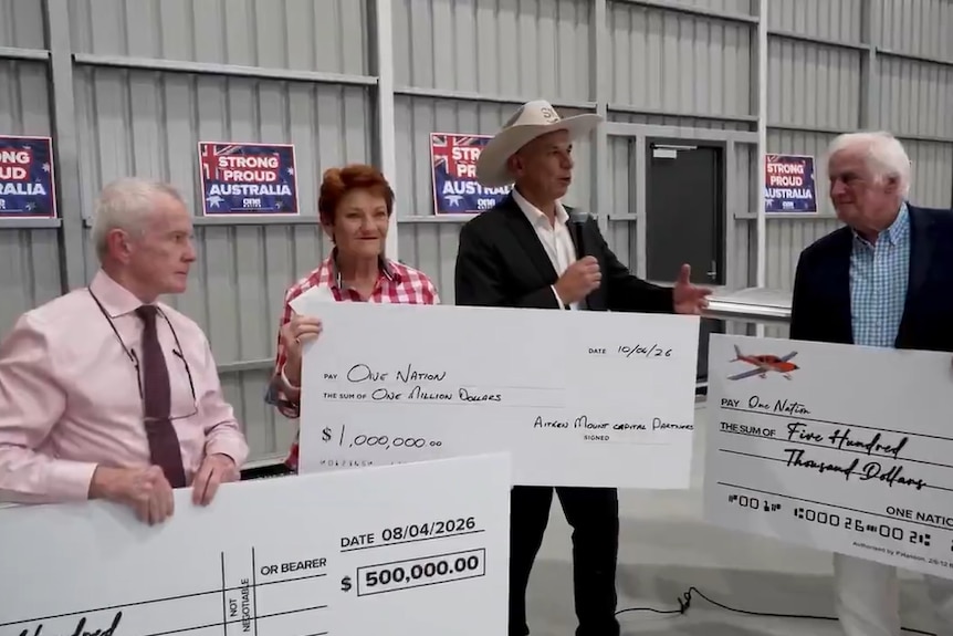 Four people stand inside a shed with three holding big novelty cheques. 
