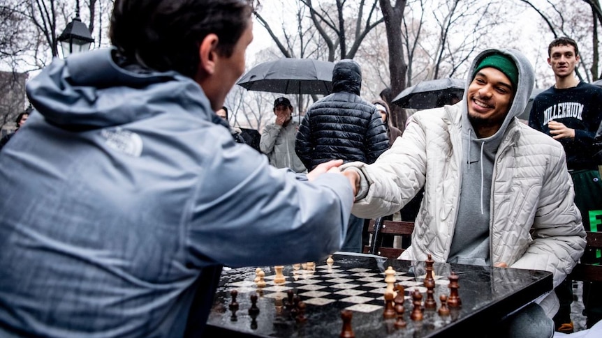 An NBA player smiles as he sits at a chess board, shaking hands with his opponent.