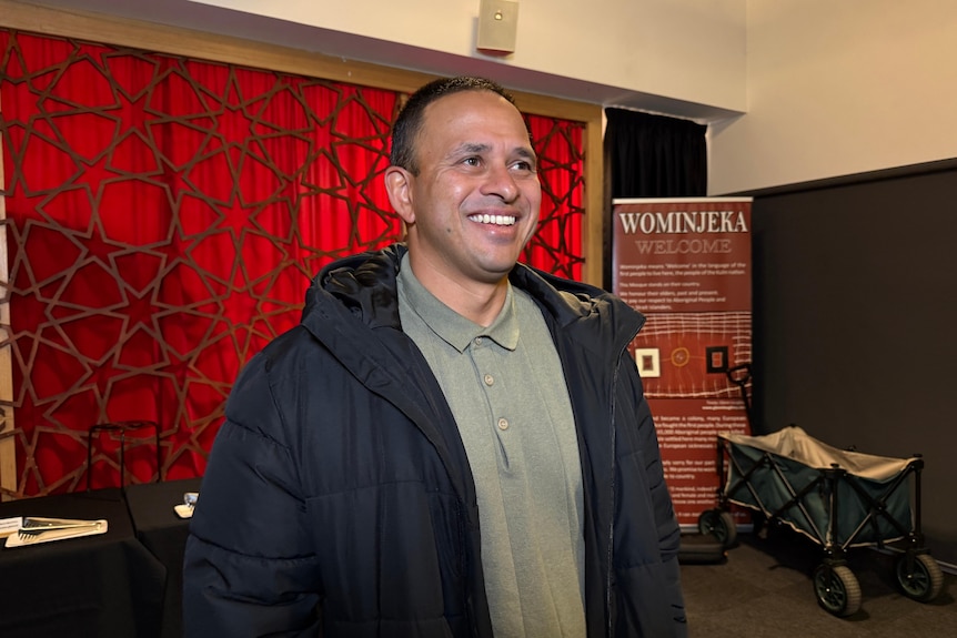 A middle-aged South Indian man in a green shirt and dark winter jacket smiles in front of red wall