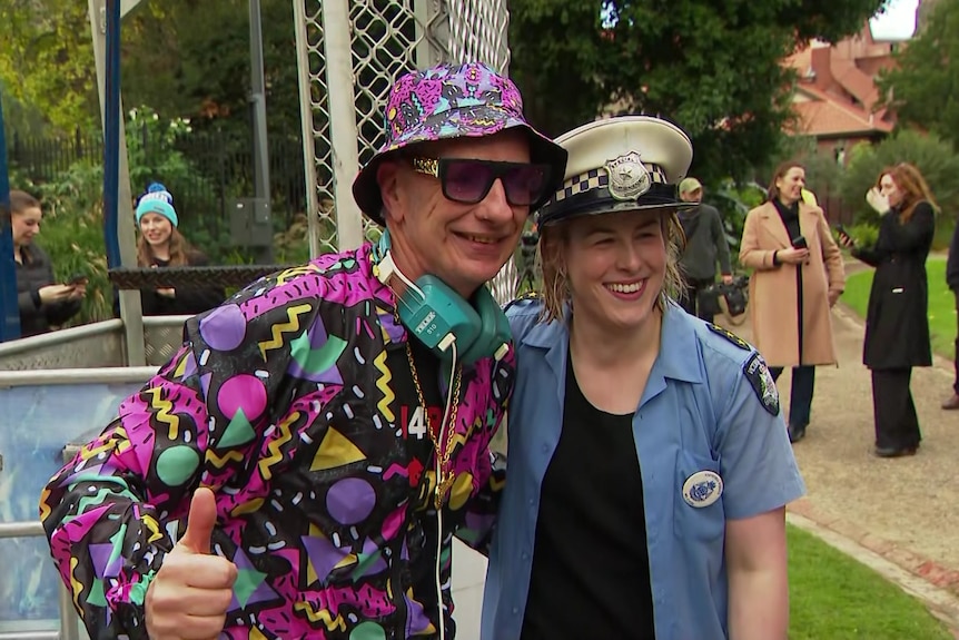 A man in a bright pink, yellow, blue and black outfit stands next to a woman dressed as a police officer.