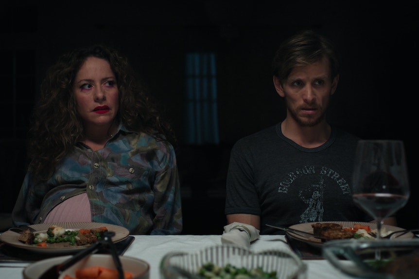 A film still of Maria Angelico and Travis Jeffery, both late 30s, looking disgusted and distressed at a dinner table.