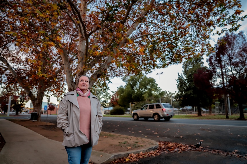 Woman wearing jumper and cream coat stands in country street imder autumn tree with car passing by