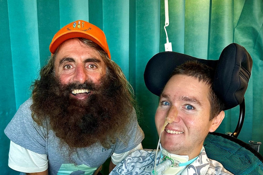 A man with a bushy beard and orange hat next to a man in a wheelchair with a feeding tube in his nostril