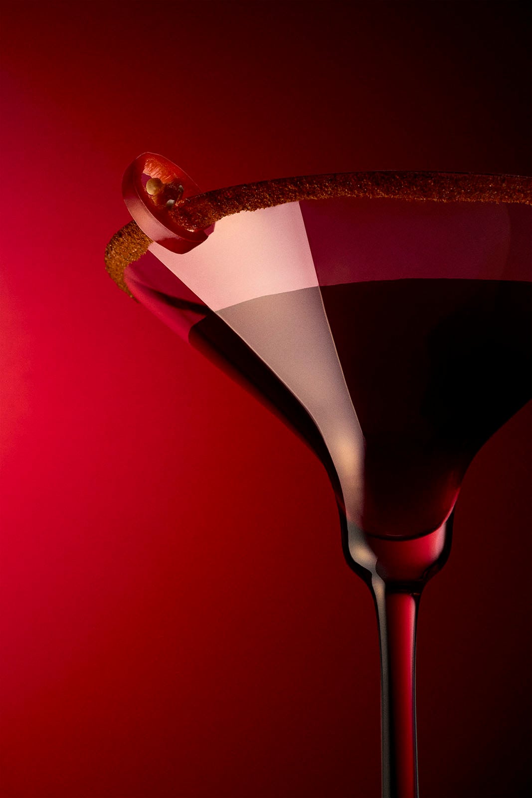 Close-up of a dark red cocktail in a martini glass with a brown sugar rim, garnished with a slice of red chili pepper, set against a rich red background.