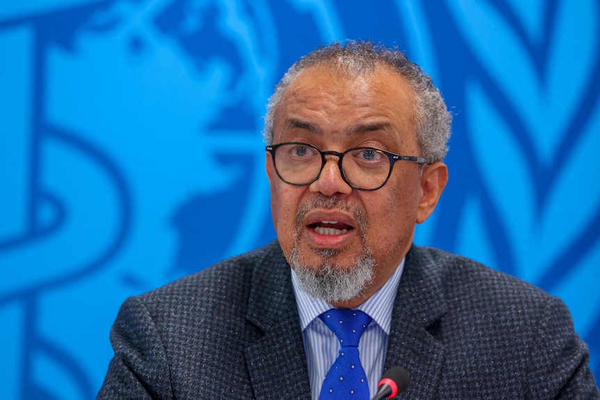 An older man with short, curly hair and glasses speaks in front of a blue backdrop.