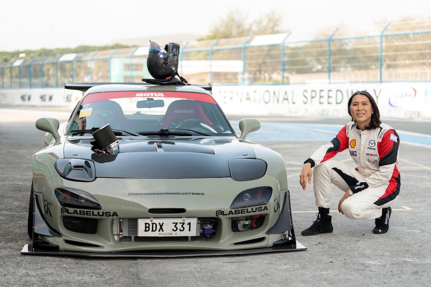 A woman crouches next to a sports car. She's wearing a race suit and is in the pits.