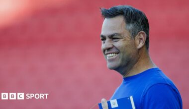 Bath director of rugby Johann van Graan smiling before a Prem Rugby mach, wearing a blue t shirt and holding a blue notebook.