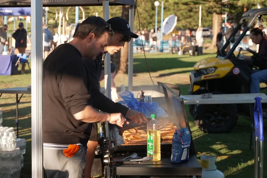 Two men use tongs to turn sausages on a bbq in a park.