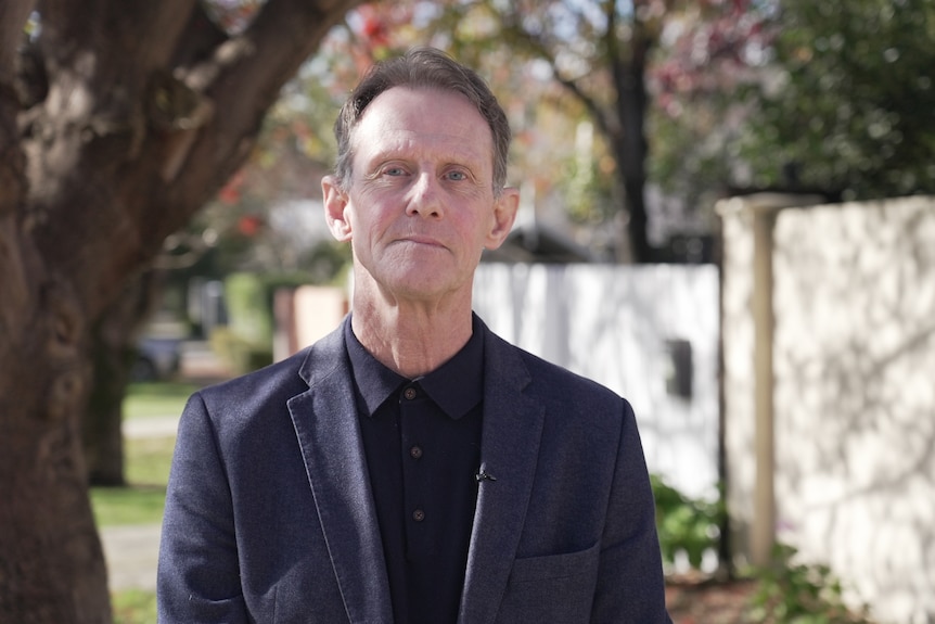 A middle-aged man wearing a dark suit and dark business shirt poses for a photo on a street outside, looking at the camera.