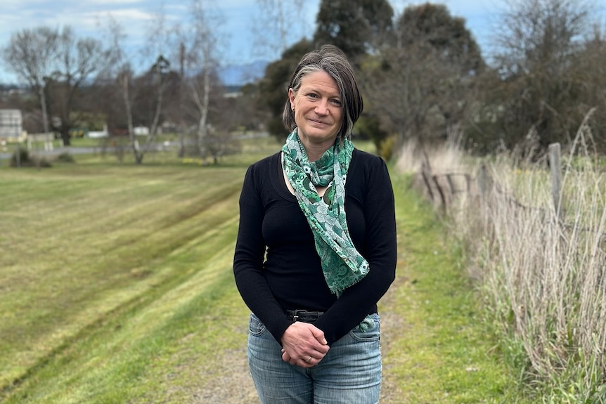 A woman with grey hair, black long sleeve shirt and a green scarf, stand in a paddock facing the camera.
