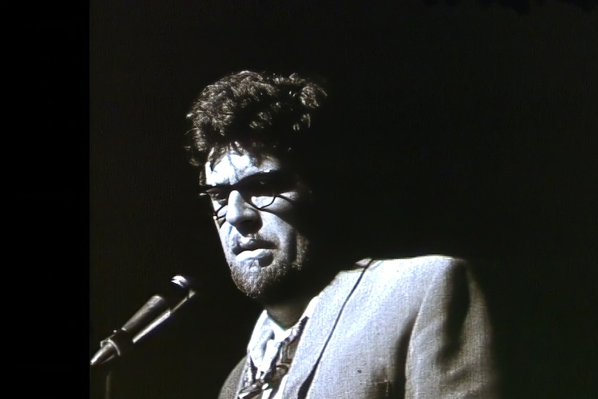 Black and white photo of a young Rolf Harris on stage by a microphone, looking sad and troubled