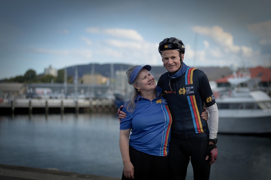 Richard Scolyer wears cycling gear, standing beside a marina, with with his arm around wife. She looks up, smiling