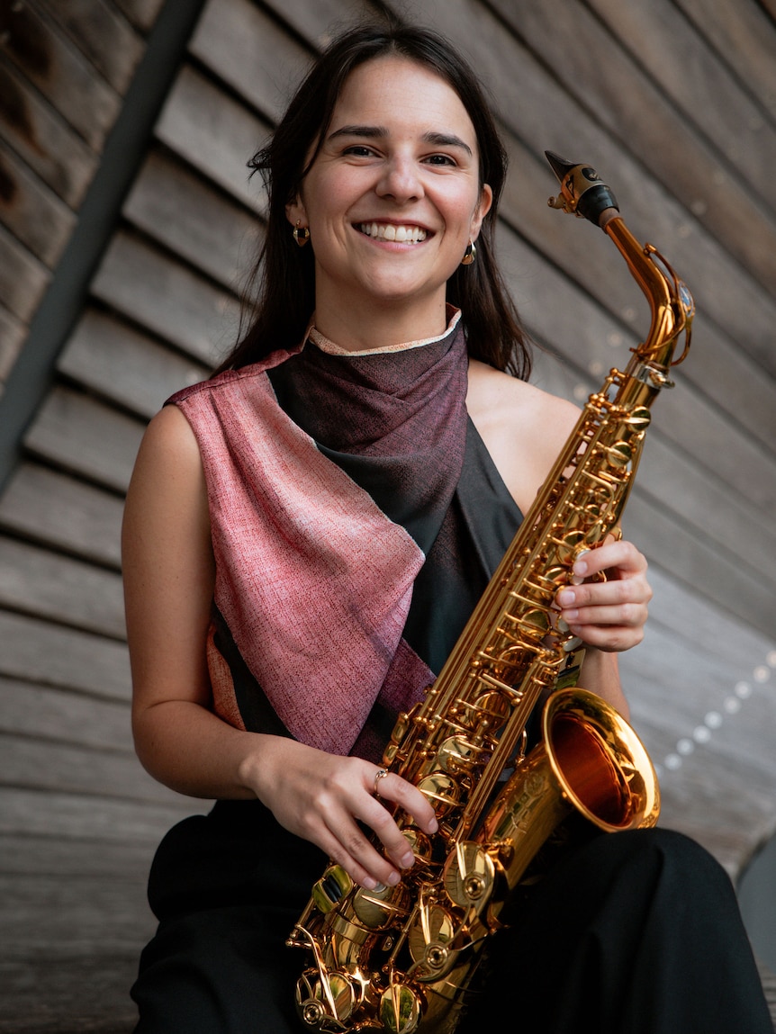 Portrait of a smiling Alice Morgan seated with a gold alto saxophone against angled wooden panels.