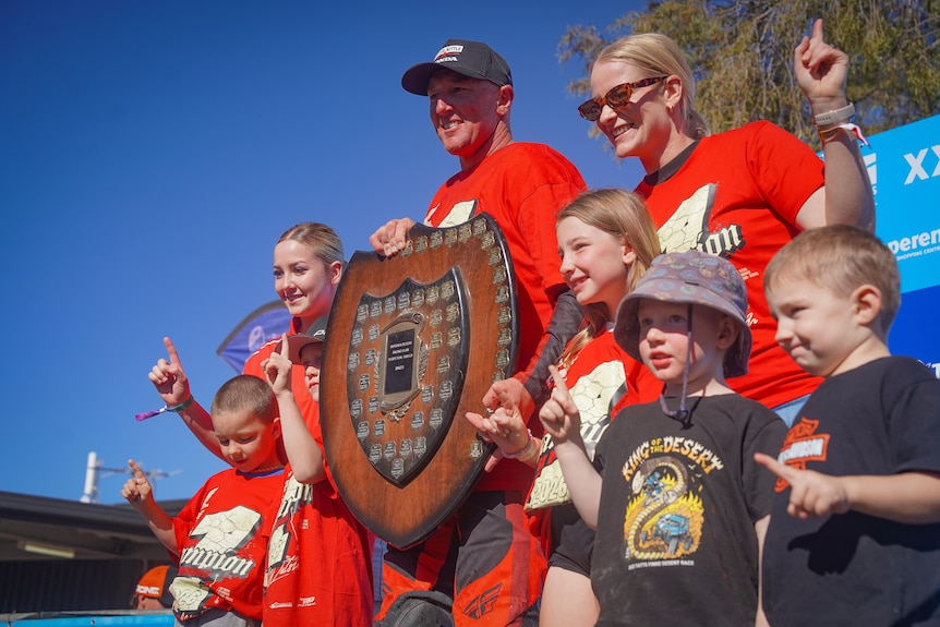 A man holding up a trophy and smiling alongside loved ones, they are all holding up a number 1 with their fingers.