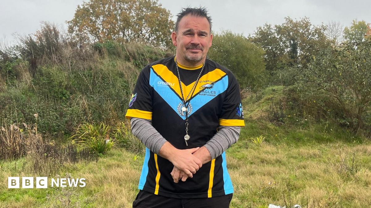 Kevin Rudd, the acting secretary of Elmbridge Eagles Rugby Club. Kevin is standing at the Oaken Lane site and is wearing training gear for the Elmbridge Eagles, including a whistle and grey undershirt.