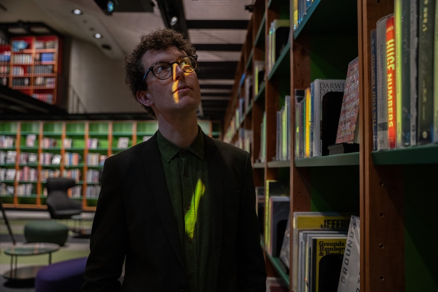 Nic Whyte gazes up at the bookshelves in a library. Colours light up the books and hit face.