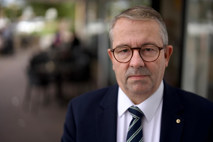 A man with glasses and in a business suit looks serious on a footpath with outdoor dining